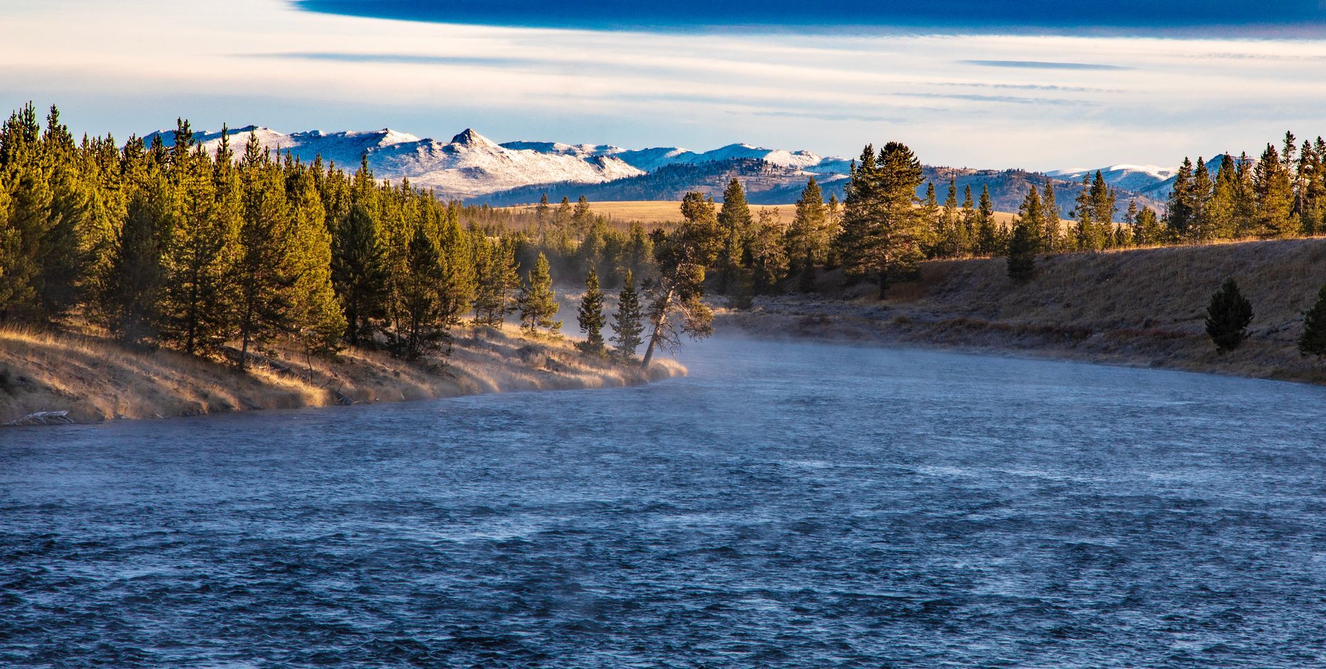 River with mist, bordered by trees and mountains in the distance; the sky is blue.