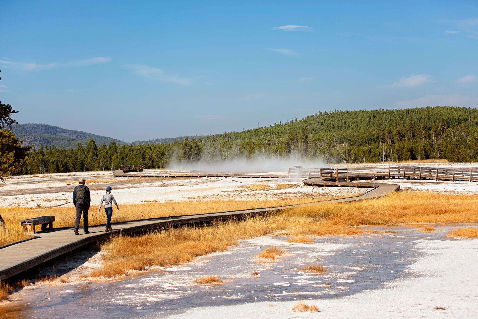 Two people walk along a wooden boardwalk through a geothermal area, with steam rising in the background.