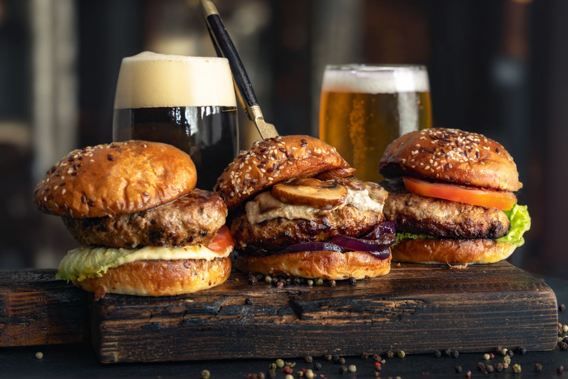 Three gourmet burgers with different toppings on a wooden board with two beer glasses.