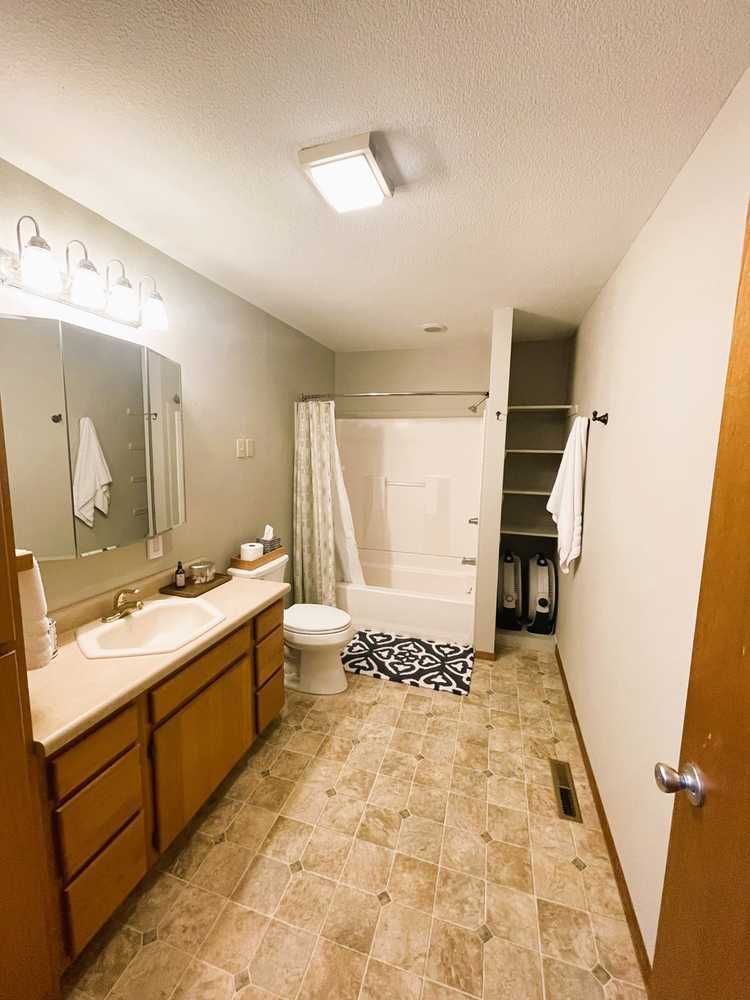 Bathroom with beige tile, white tub, long wood vanity, and a tall shelf.