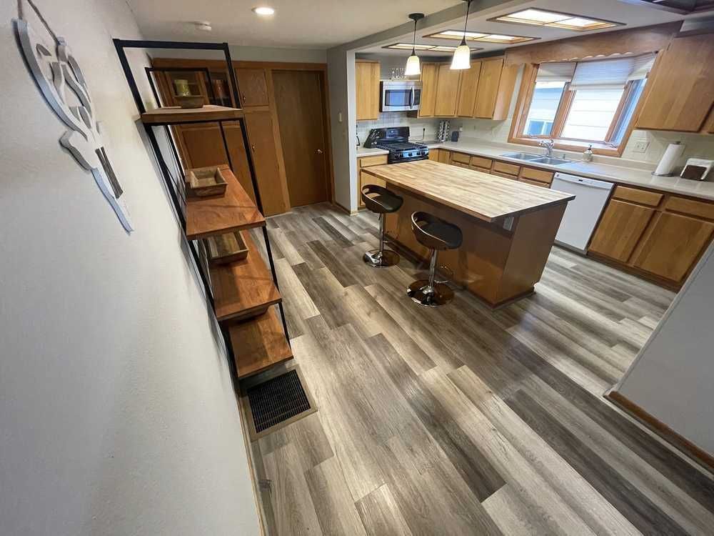 Kitchen with wooden cabinets, island with stools, and wood-look flooring. A shelving unit is on the wall.