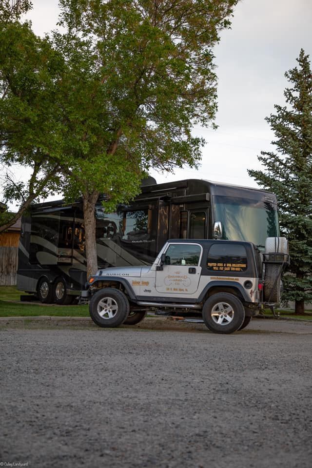 Silver Jeep in front of a large black RV parked on gravel. A tree and evergreens are in the background.