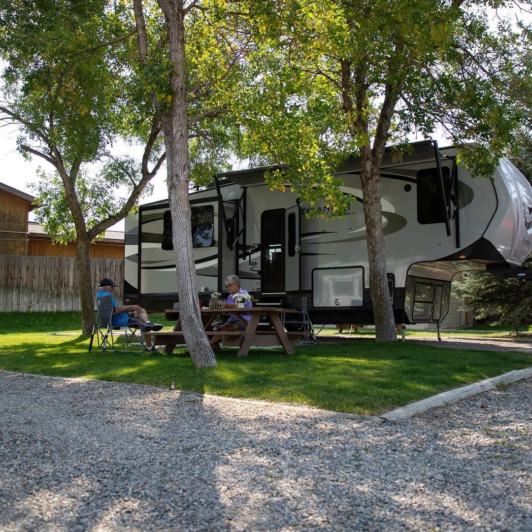 RV parked in a campground, people at picnic table and in chairs on the grass. Trees provide shade.