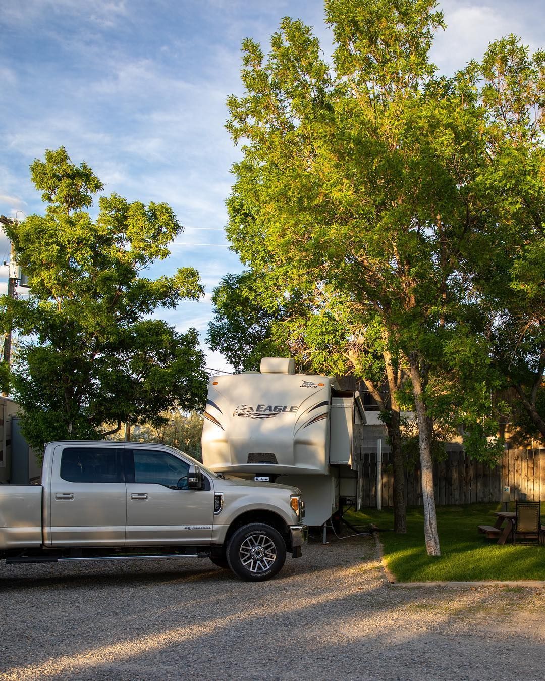 Silver pickup truck parked next to a camper under a tree, gravel ground, blue sky.