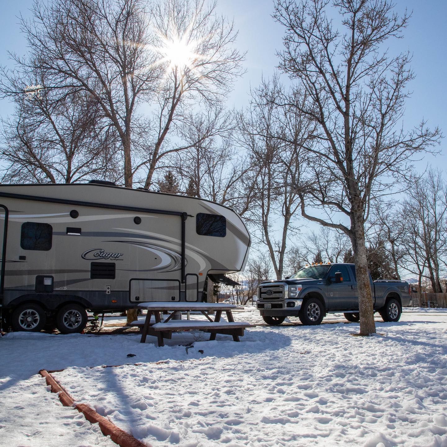 RV and truck parked in snowy campground, picnic table in front. Sun shining through bare trees.