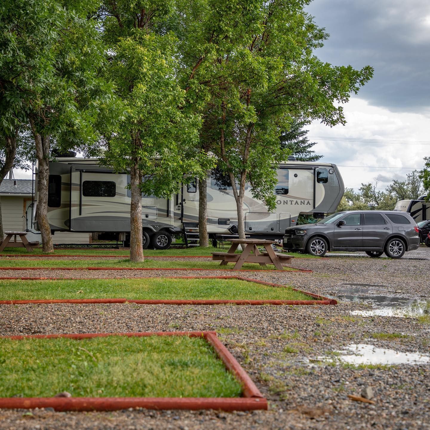 RV campground with campers, picnic table, trees, and gray SUV under cloudy sky.