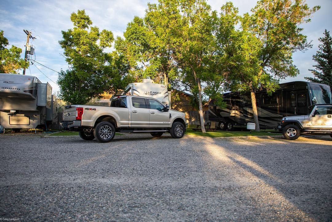 A light-colored pickup truck parked on gravel at a campsite, surrounded by trees and RVs.