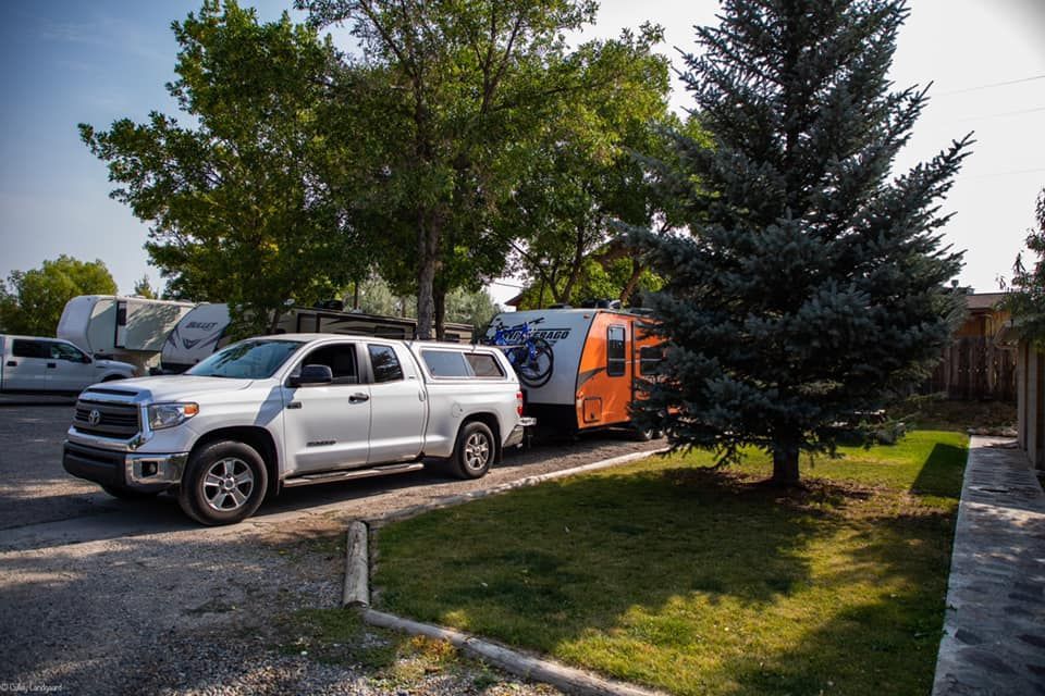 White pickup truck towing a small orange and blue camper, parked on gravel next to grass and trees.