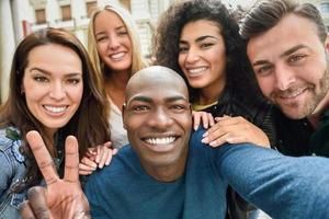 Cinco pessoas sorridentes tiram uma selfie ao ar livre.