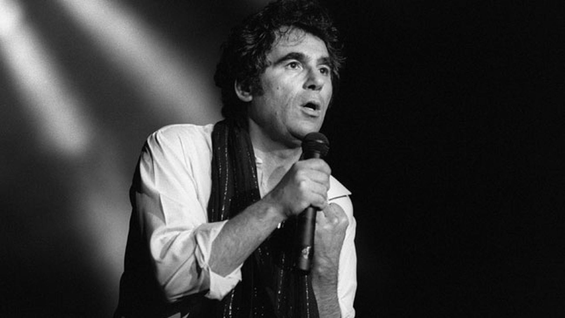 Een man zingt in een microfoon op een podium op een zwart-witfoto.