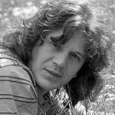 Een zwart-witfoto van een man met lang krullend haar die in het gras ligt.