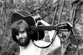 Een man met een baard houdt een gitaar over zijn schouder op een zwart-witfoto.