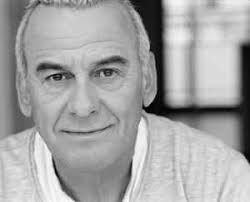 Een zwart-witfoto van een oudere man die naar de camera lacht.