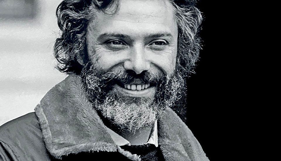 Een man met een baard en krullend haar lacht op een zwart-witfoto.