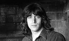 Een zwart-witfoto van een man met lang haar die voor een bakstenen muur staat.