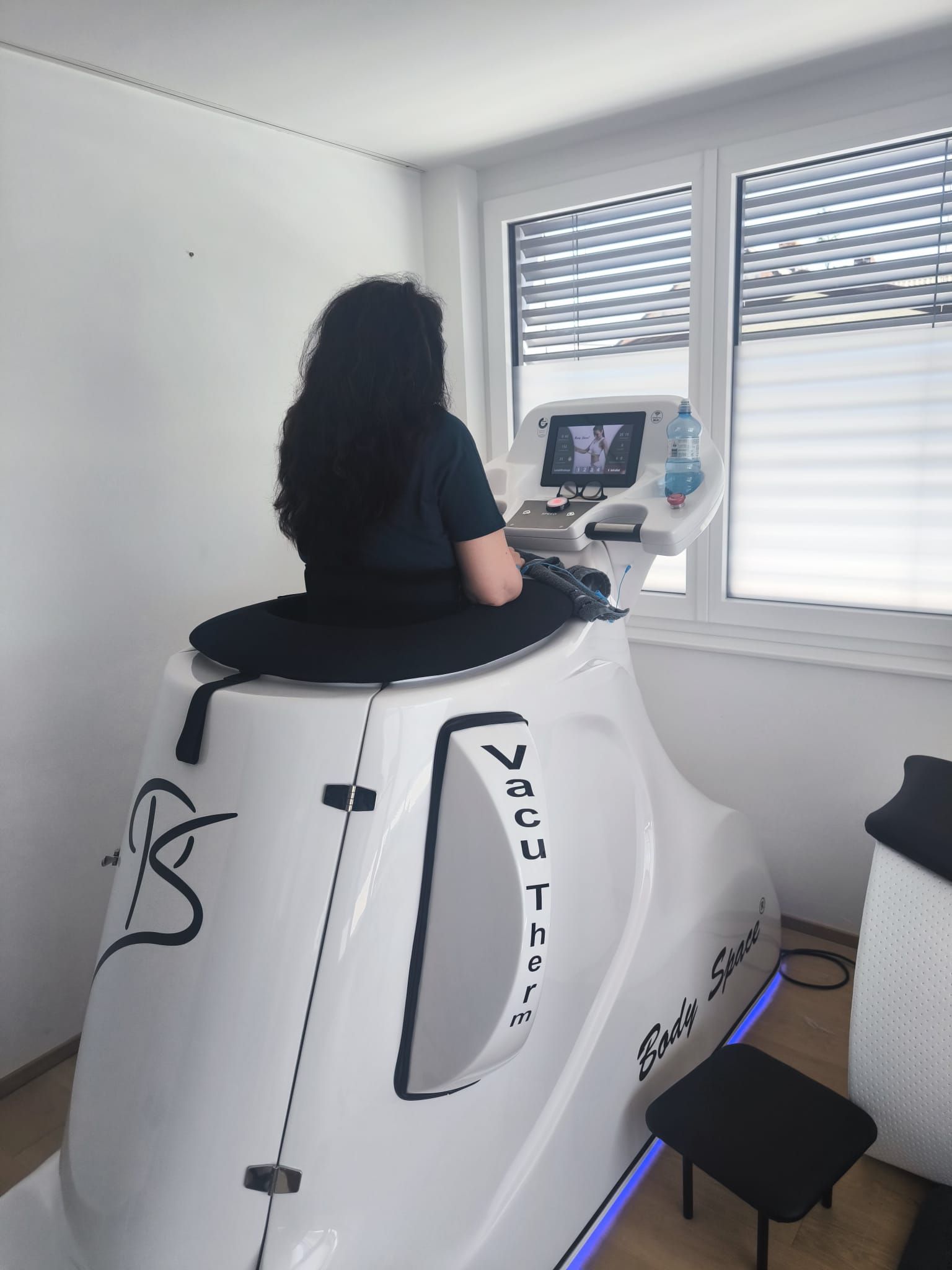 Frau sitzt auf einem weißen VacuTherm-Fitnessgerät in der Nähe eines Fensters. Ihr Blick blickt auf das Bedienfeld.