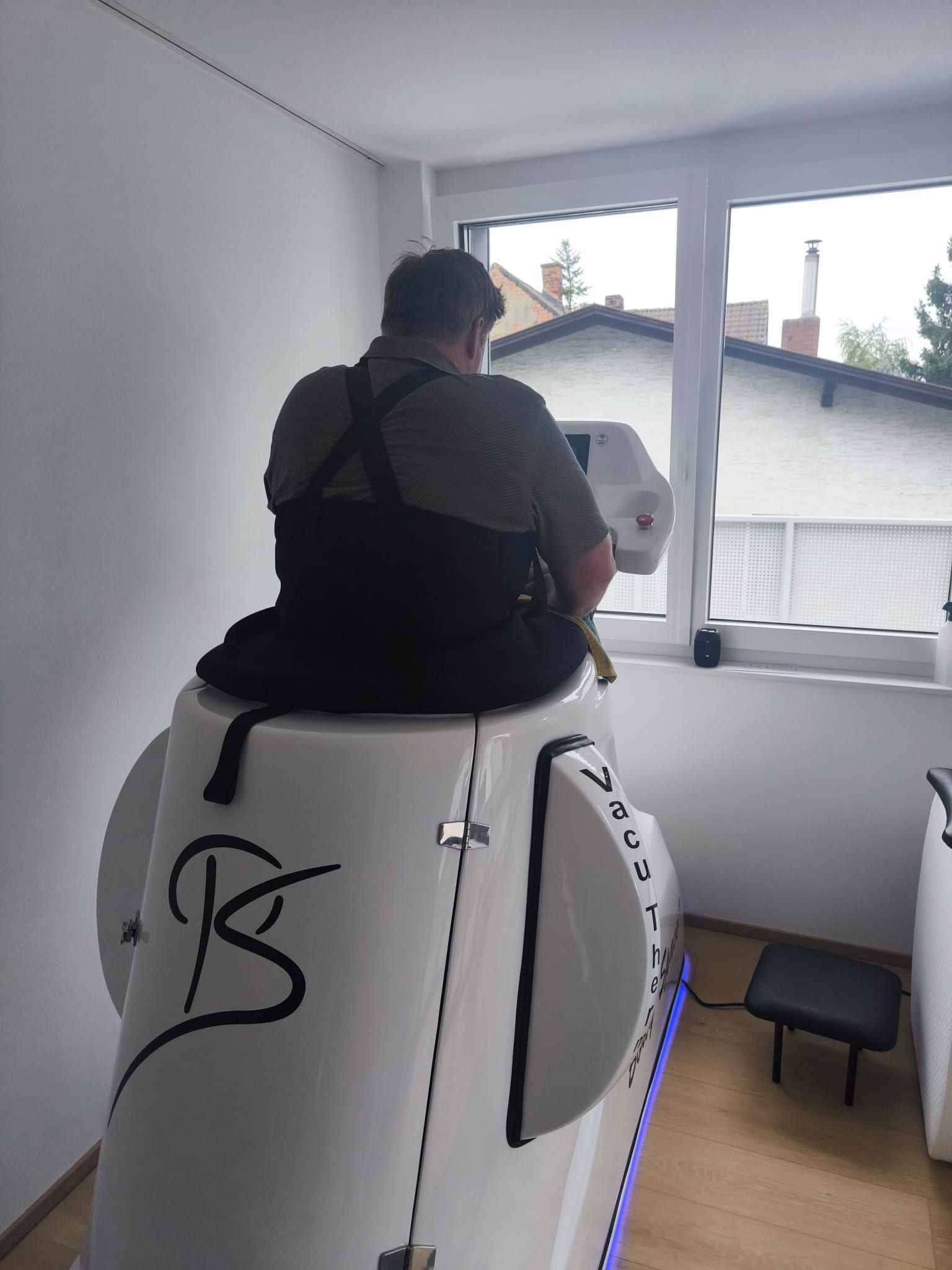 Ein Mann sitzt in einem weißen Trainingsgerät mit dem Logo „BS“. Er blickt auf einen Bildschirm und aus dem Fenster.