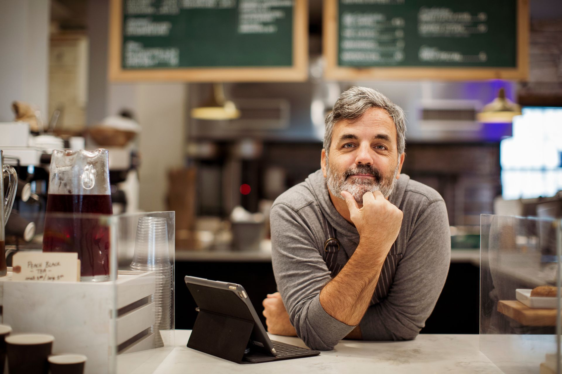 Man in a cafe, smiling, resting chin on hand, near counter with tablet and drinks, with menu behind.