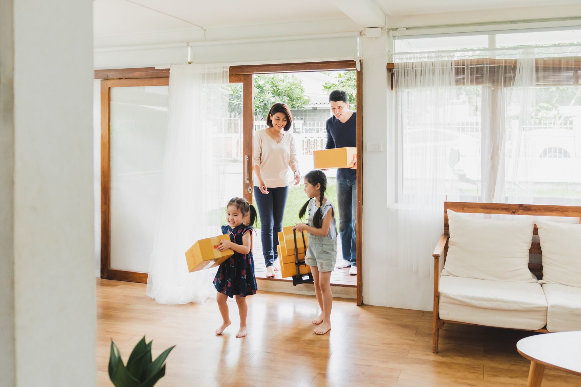 Family entering a new home carrying boxes. Two young girls lead, parents follow. Bright, sunny doorway.