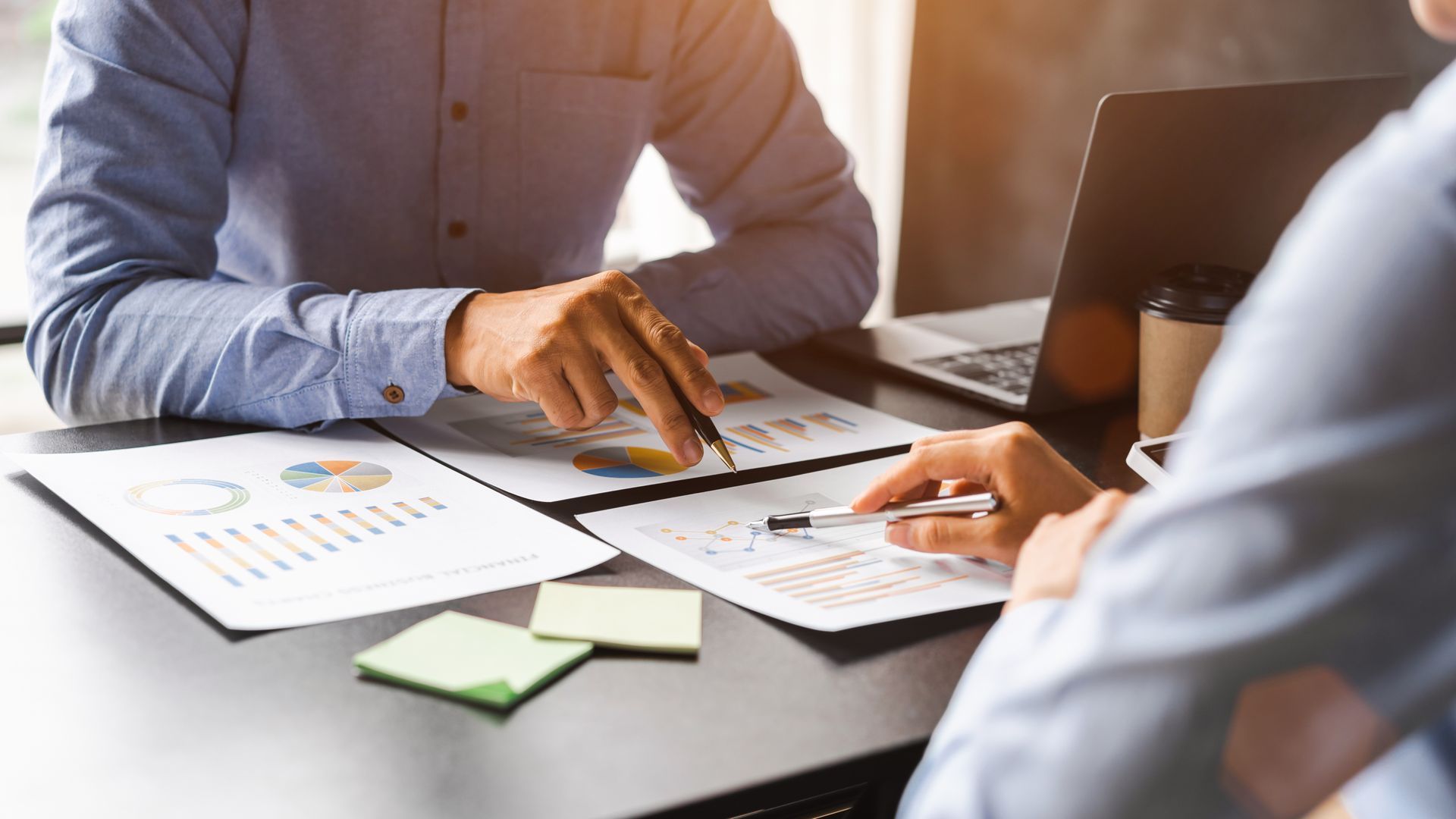 Two people in a business meeting, reviewing financial documents with pens and a laptop.
