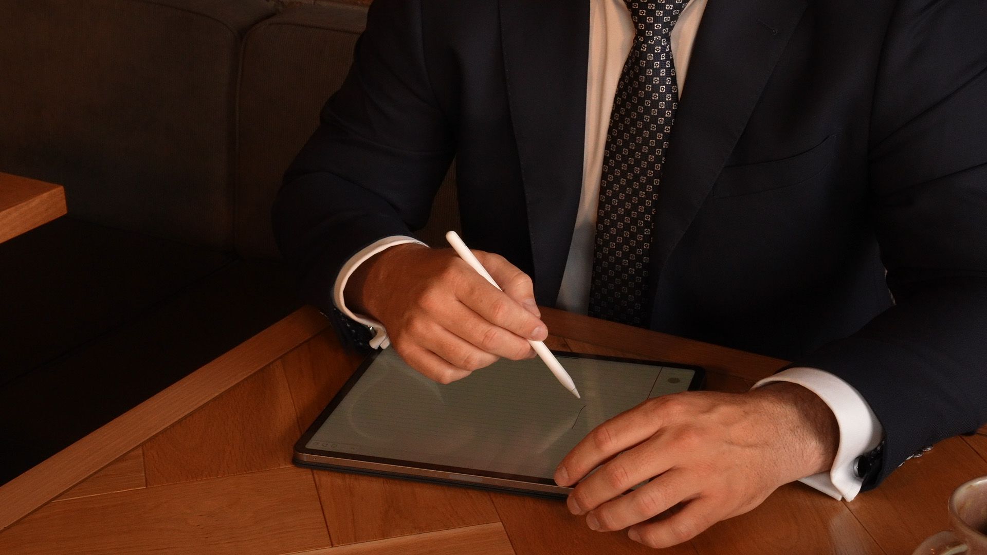 Person in a suit using a stylus on a tablet at a wooden table.