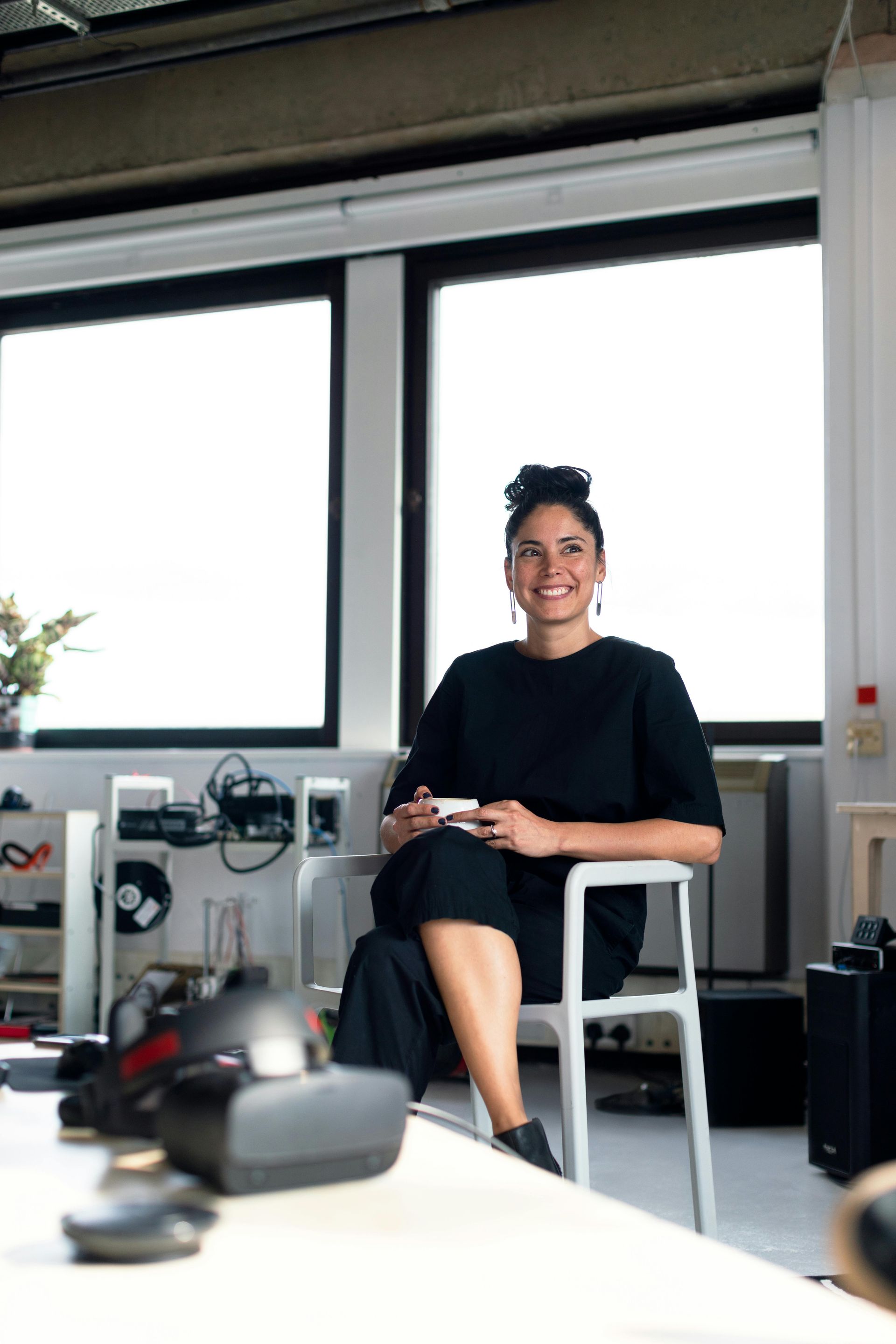 Woman in black dress sits smiling, holding mug, near windows, VR headset visible.