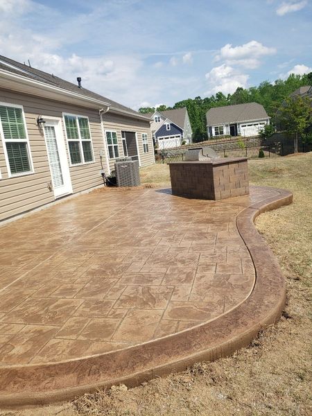 Stamped concrete patio beside a brick house, with a curved edge and outdoor kitchen island in a backyard.