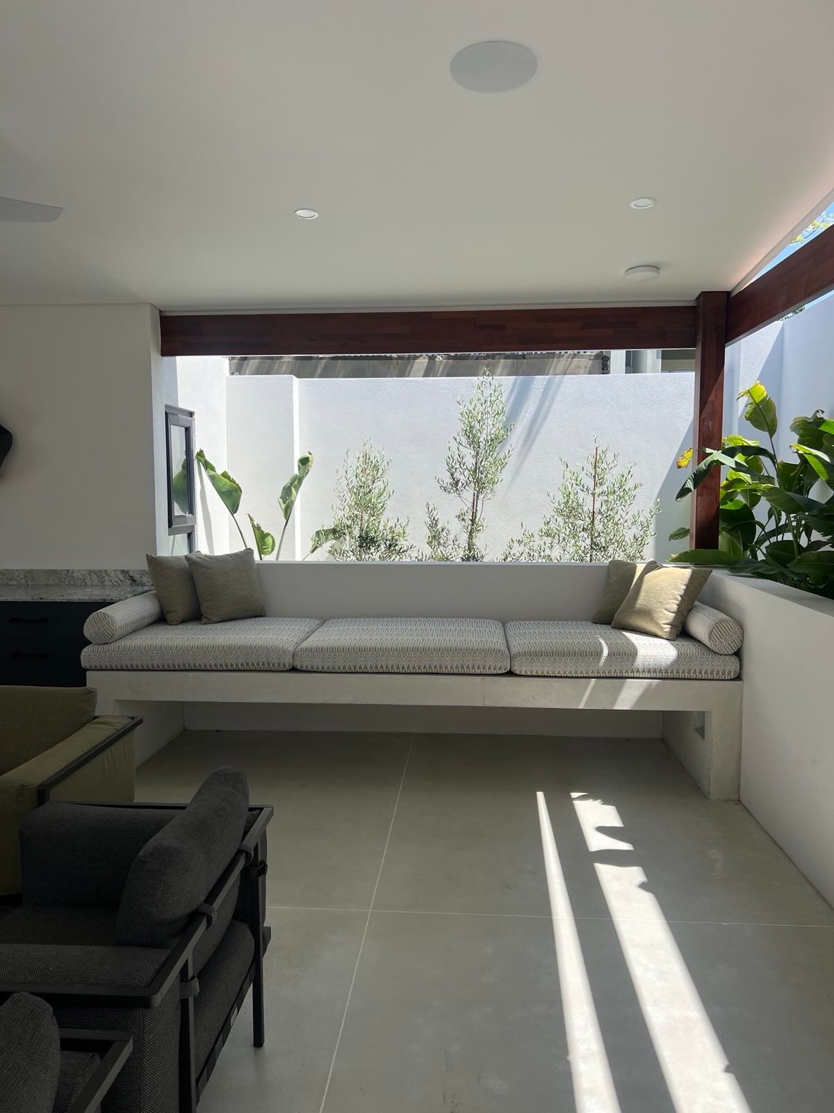 Built-in concrete outdoor sofa with cream cushions and pillows, looking out to greenery, in a white-walled space.