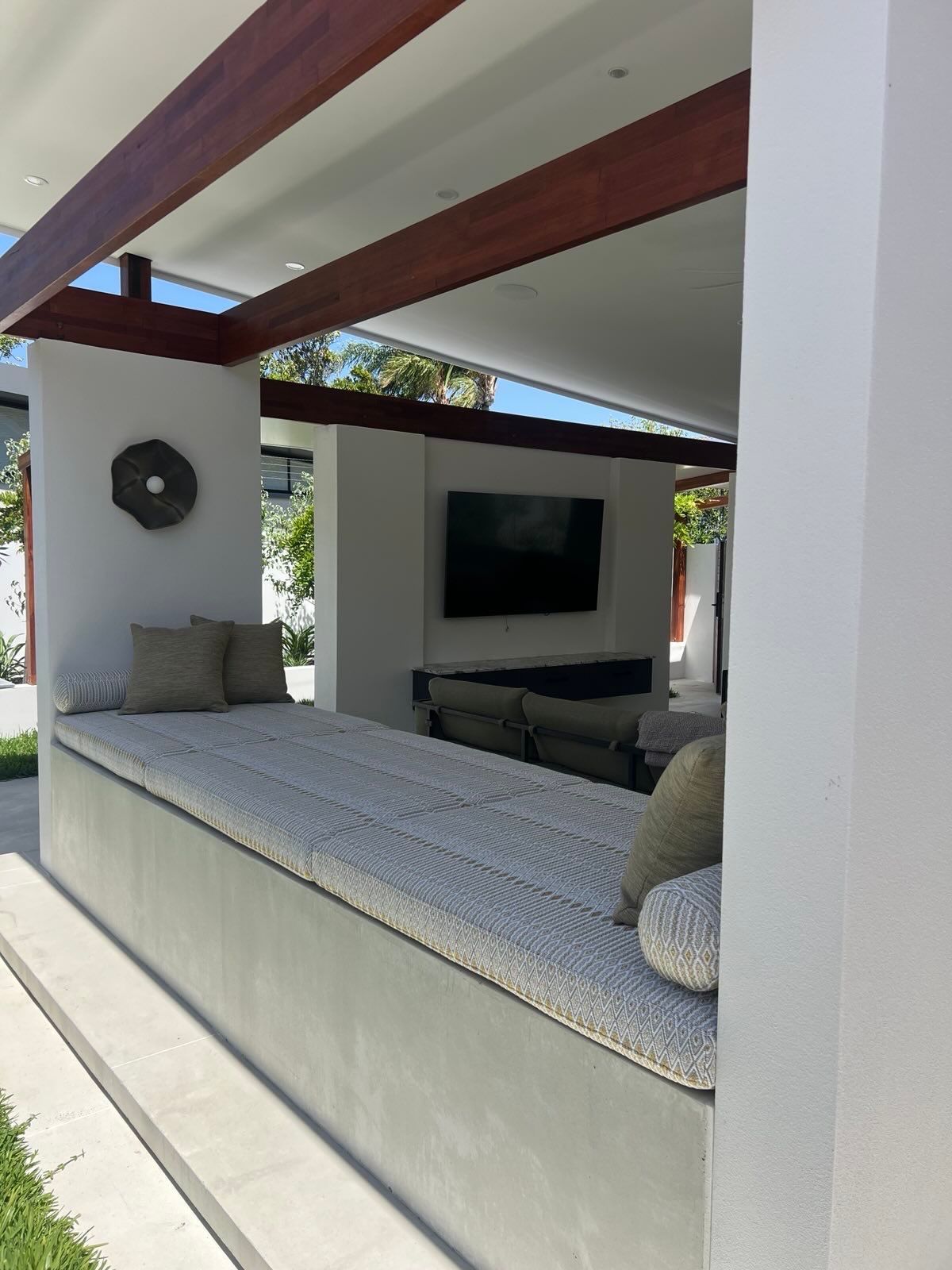 Outdoor lounge area with built-in concrete seating and day bed, cushions, TV, and wooden beams.