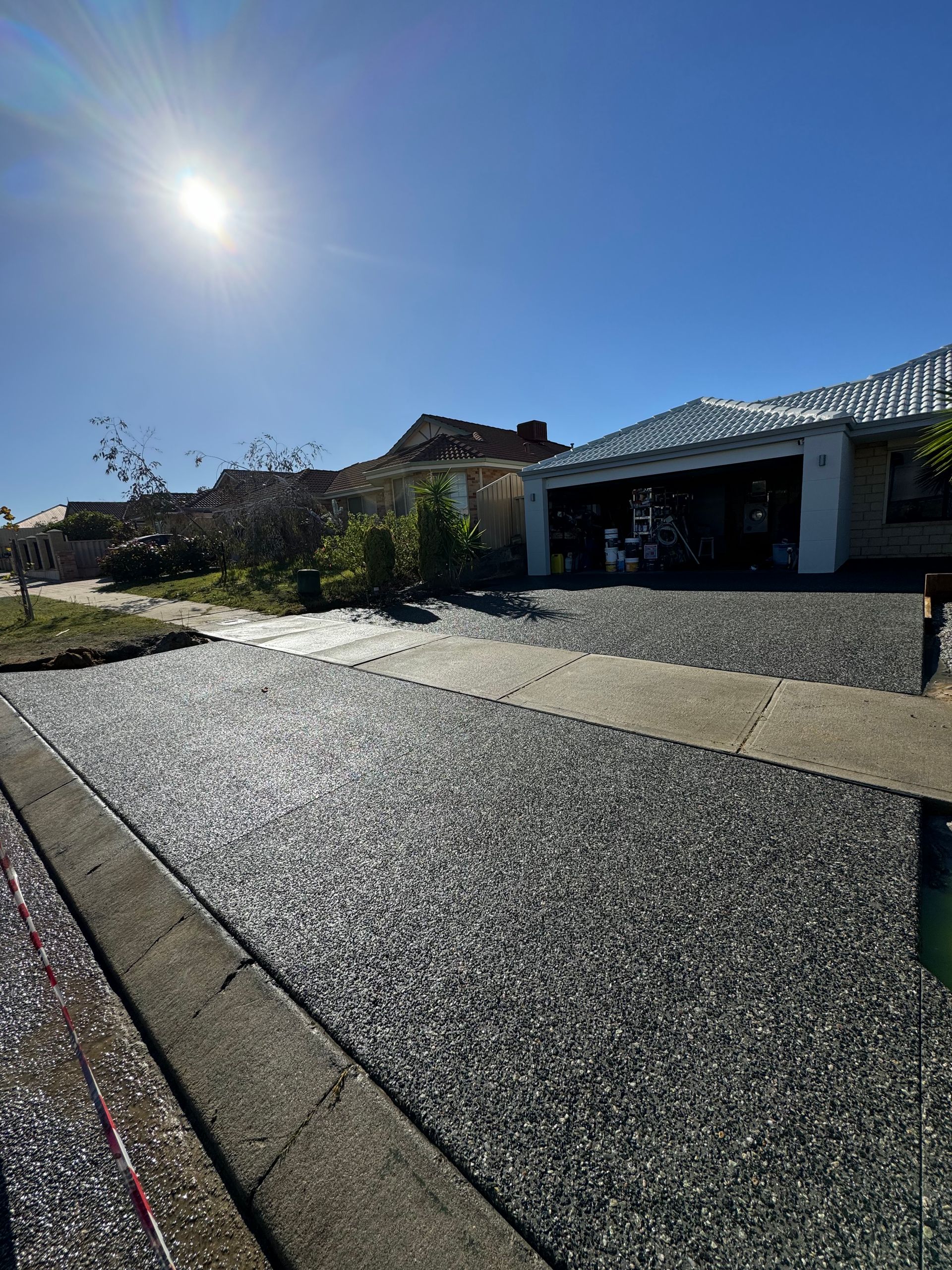 Driveway with black exposed aggregate surface leading to a house, bright sun in the sky.