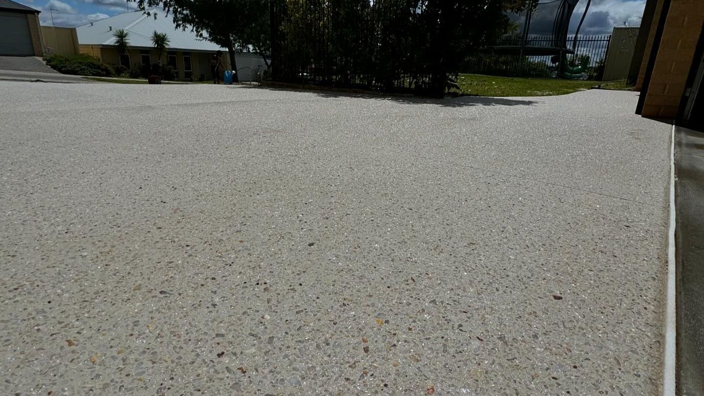 Driveway with exposed aggregate and surrounding it is green grass and gardens