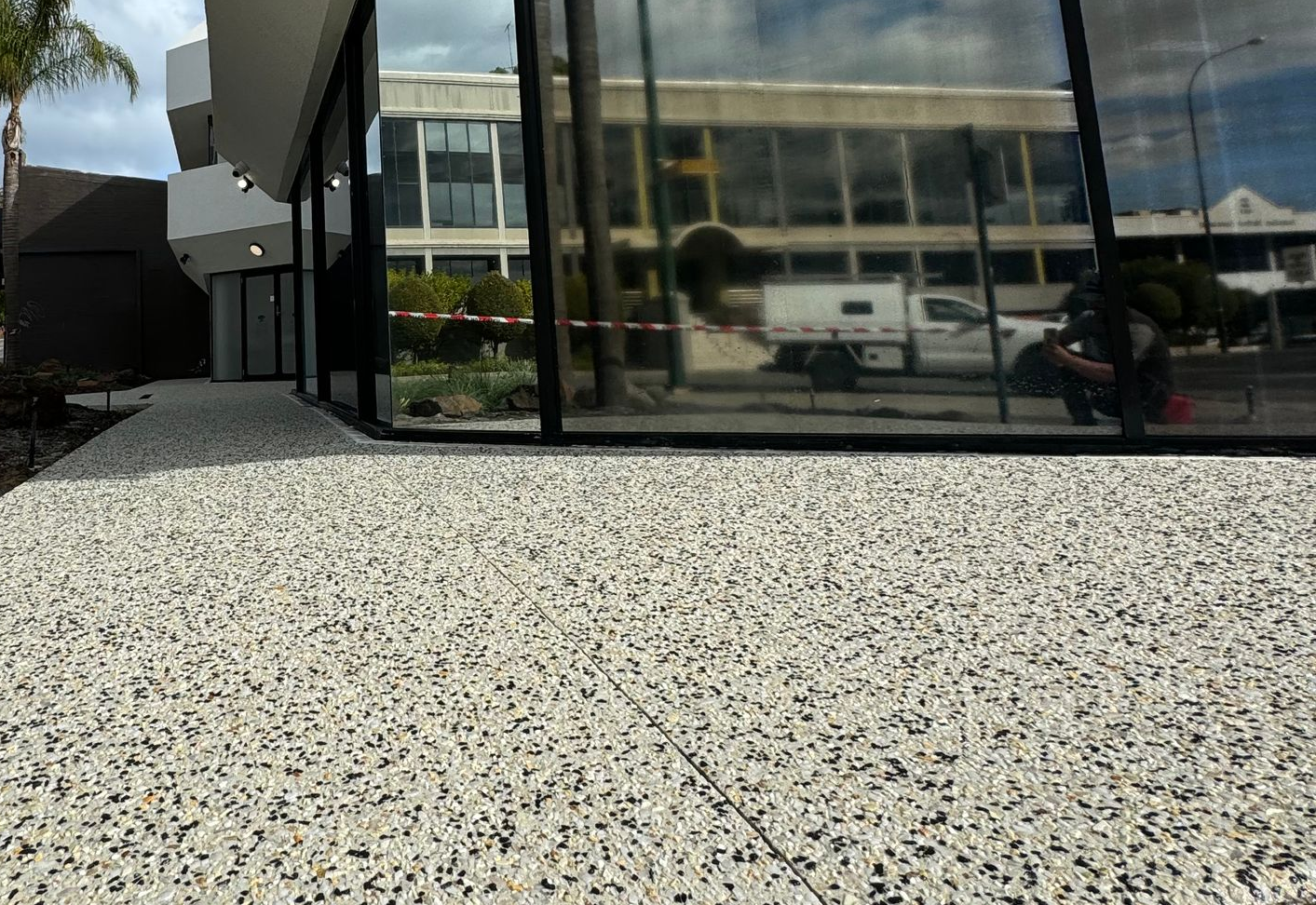 A modern building exterior featuring a textured, white and black speckled concrete pathway and glass windows reflecting the street.