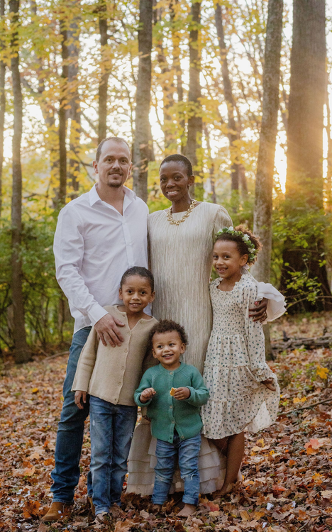 A family of five poses for a portrait in a sunlit, wooded area filled with fallen autumn leaves.