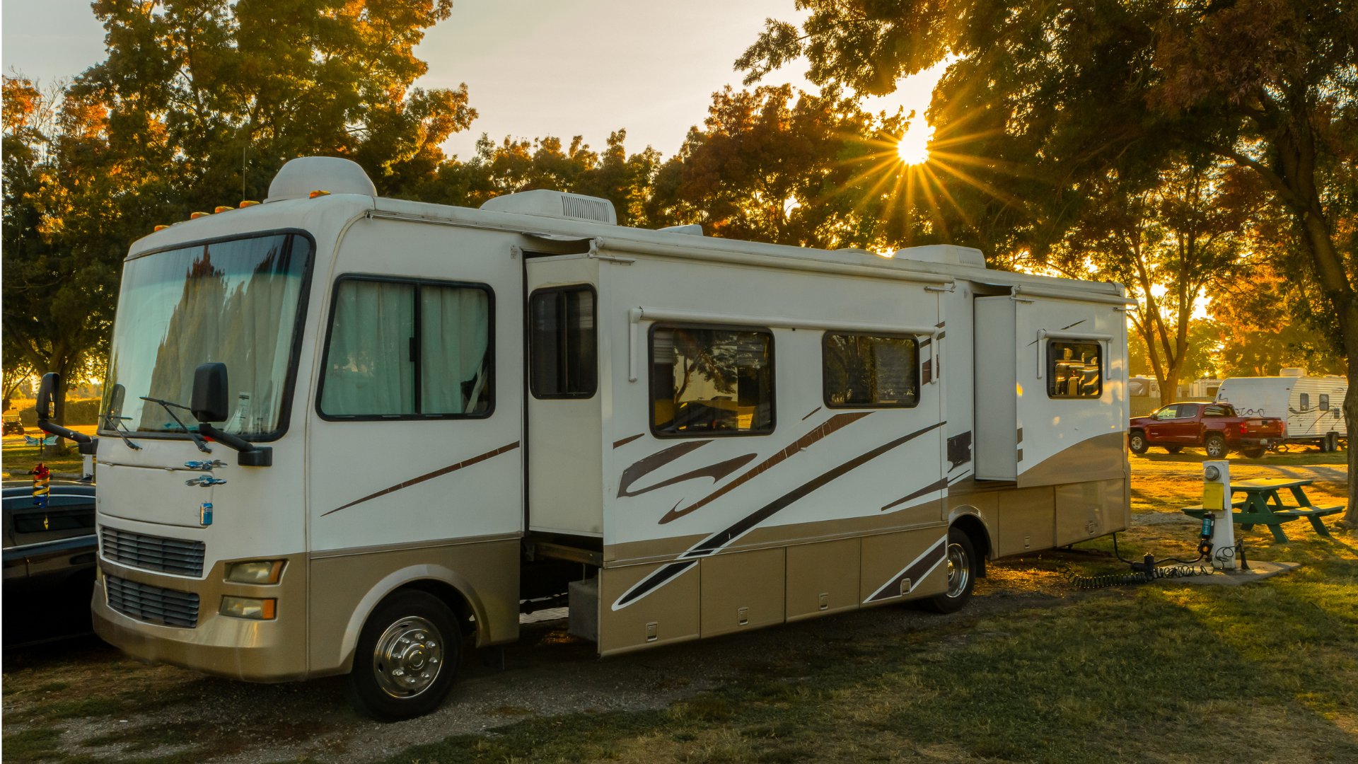 A vintage white and brown RV parked on a tree-lined street covered in fallen autumn leaves, with a canoe on its roof.