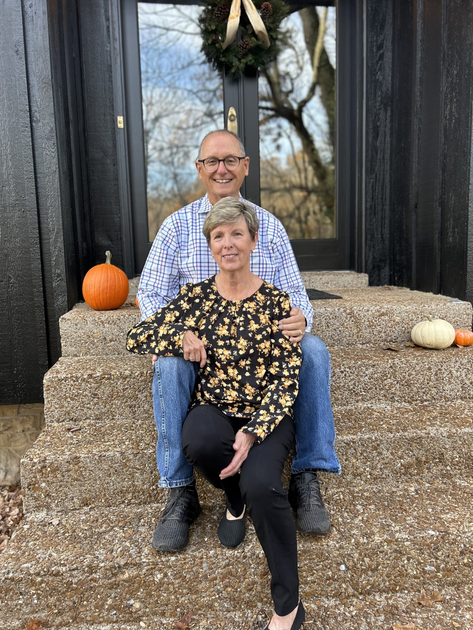 A couple sits smiling on the steps of a porch in front of a dark door, flanked by orange and white pumpkins.