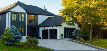 Maison moderne à deux étages avec toit gris, bardage blanc et panneaux décoratifs foncés. Une allée pavée mène au garage, avec un grand arbre et des buissons dans le jardin.