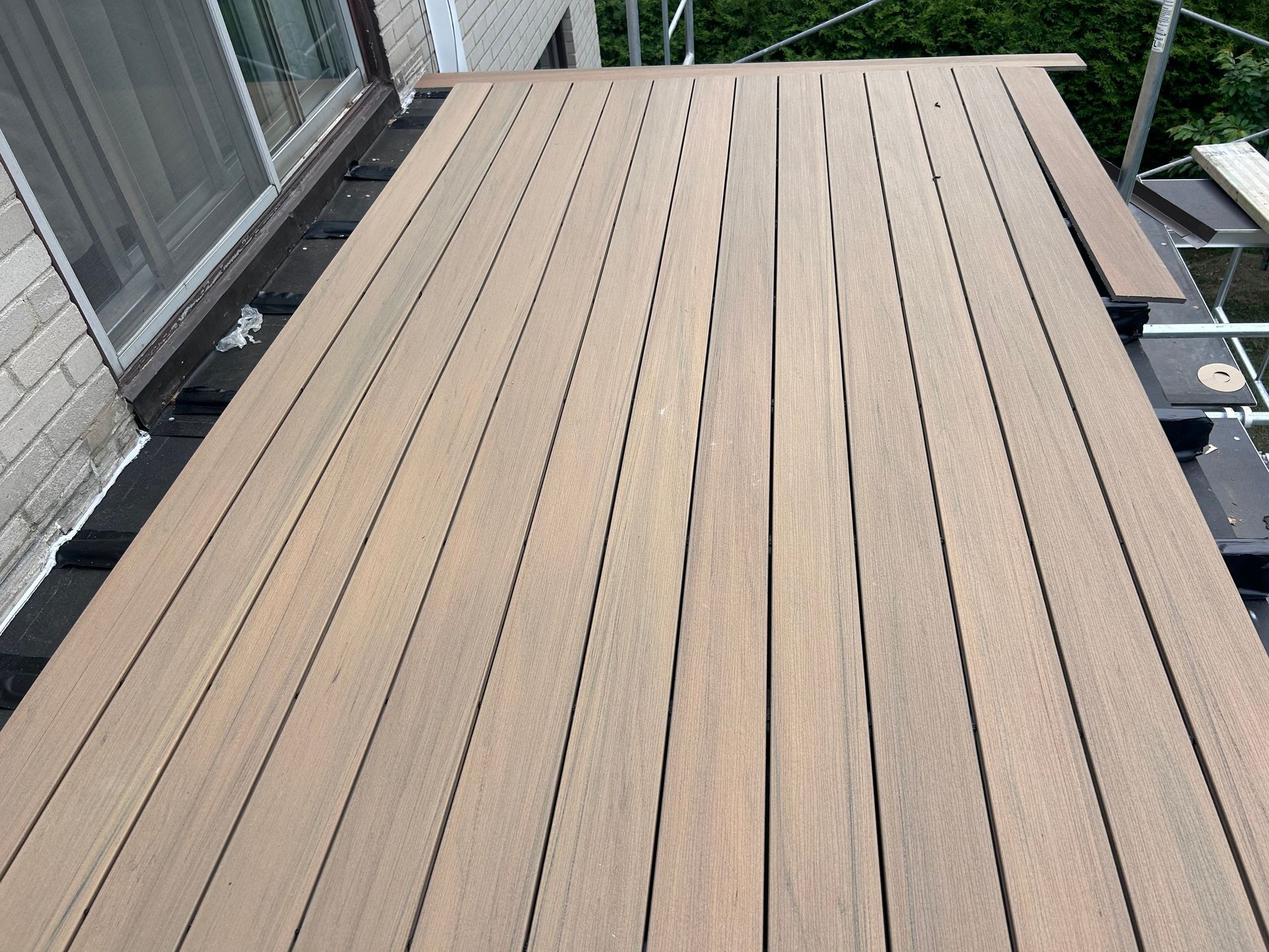 Planches de terrasse en bois brun clair disposées à côté d'une porte coulissante en verre. Construction en cours, outils et échafaudages visibles.