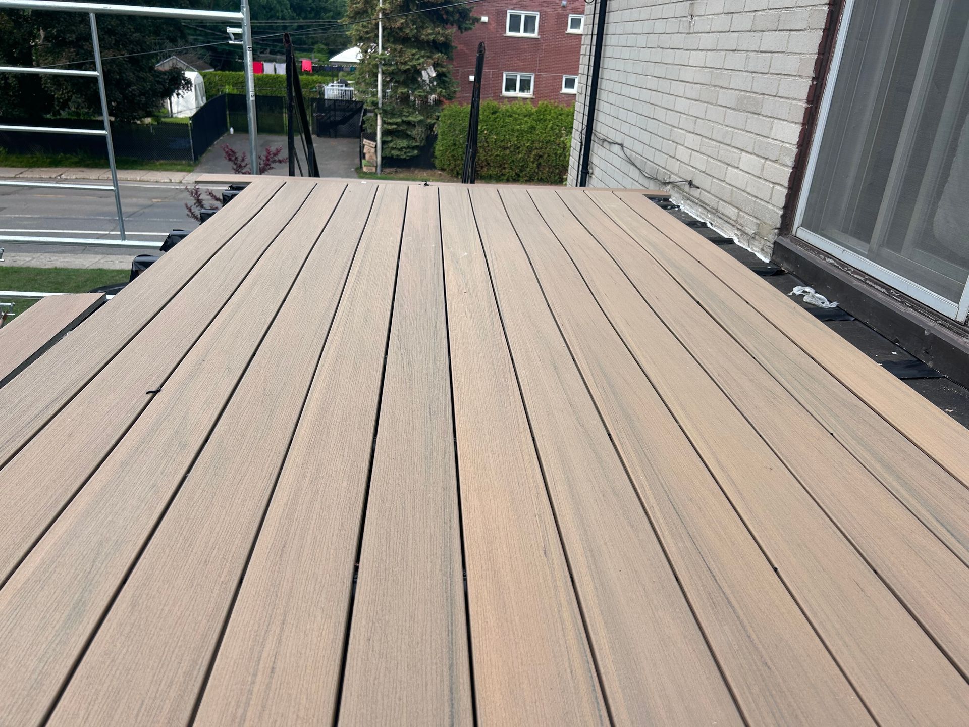 Vue aérienne d'une terrasse en planches de bois clair. La terrasse est située à côté d'un bâtiment et donne sur une rue avec une clôture et des arbres en arrière-plan.