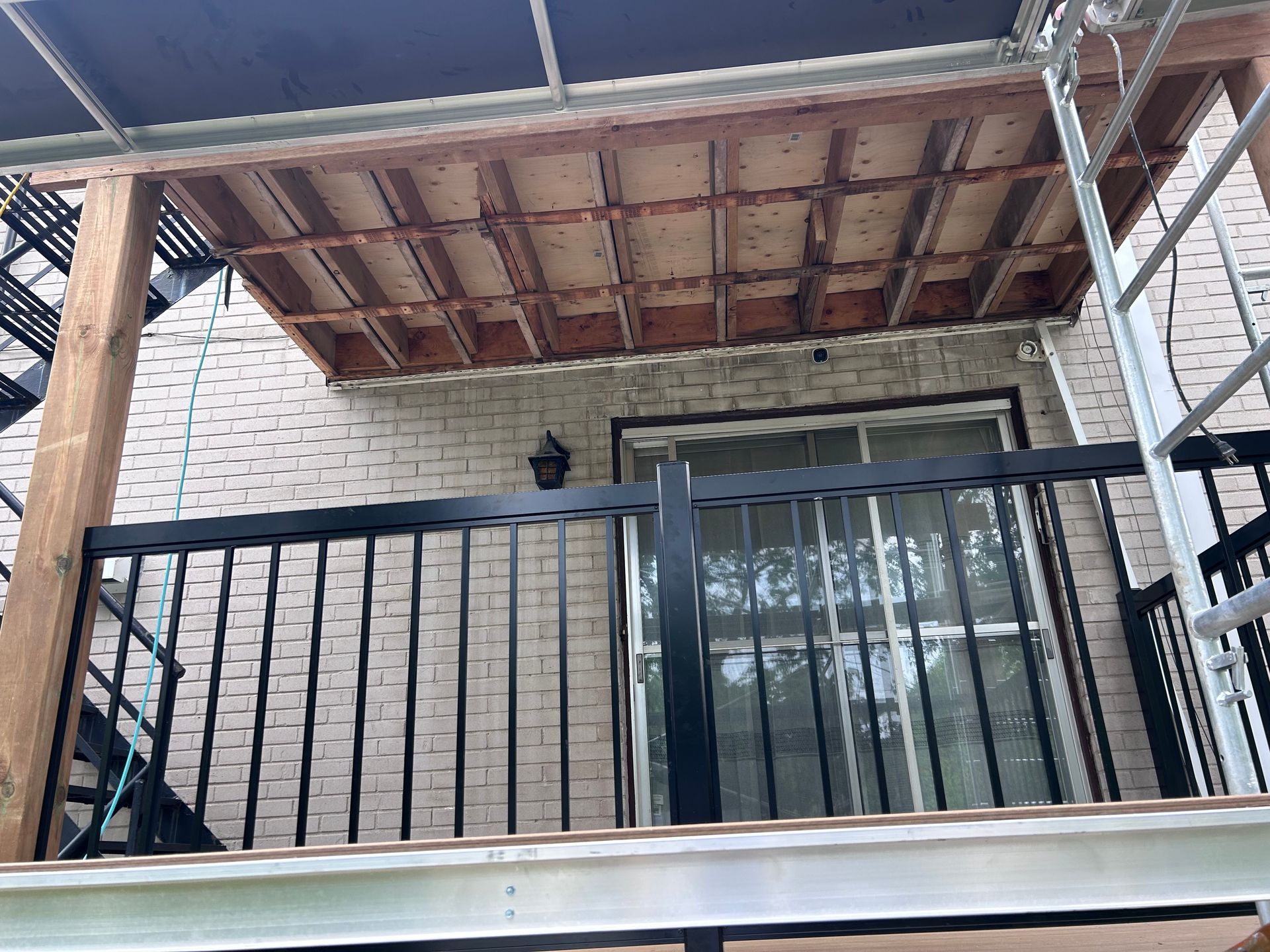 Balcon partiellement construit, attenant à un bâtiment en briques. On peut voir une balustrade noire et un plafond en bois brut.