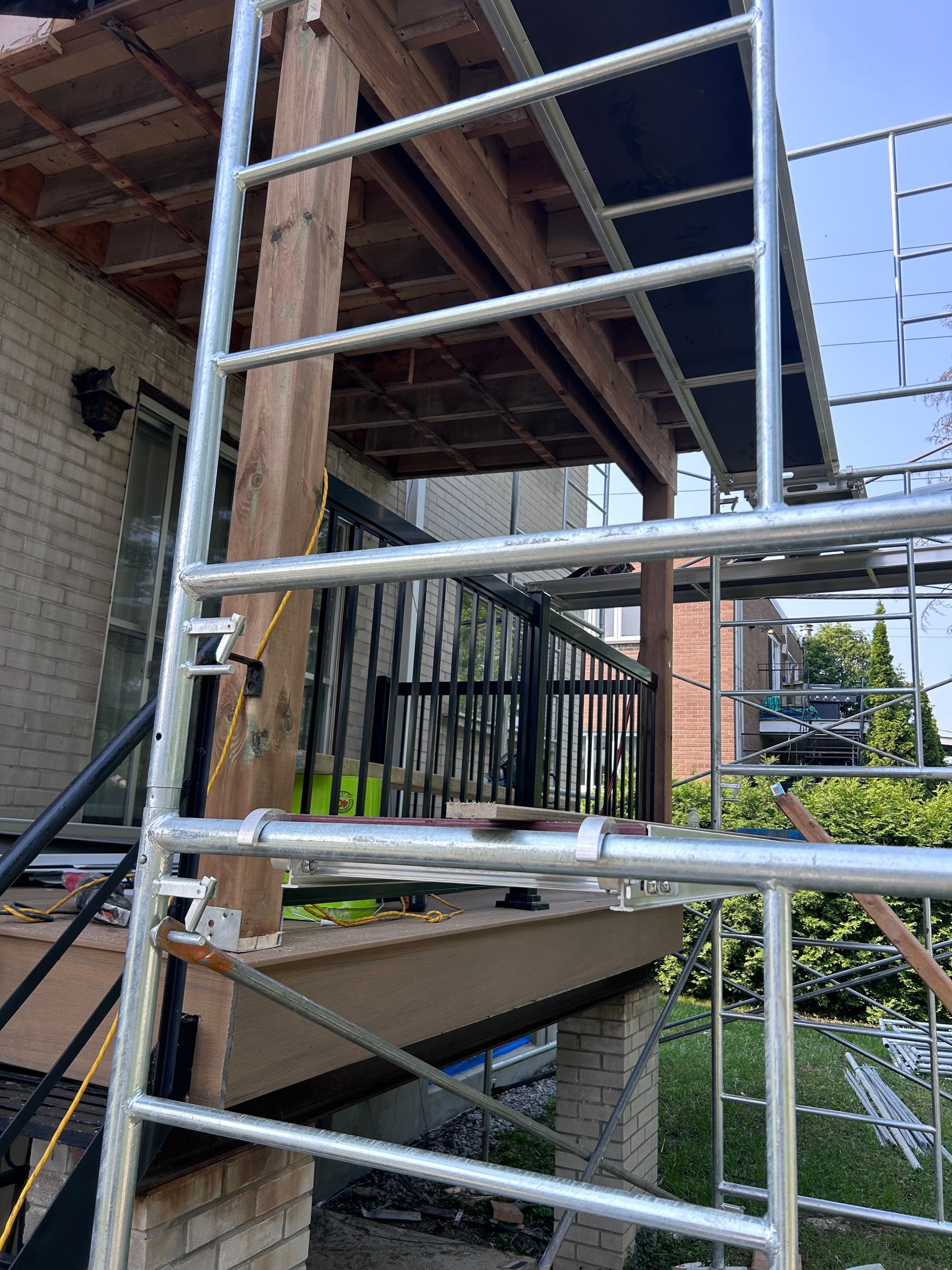 Un échafaudage métallique à côté d'un porche avec une balustrade noire, des poutres en bois marron et des piliers en briques.