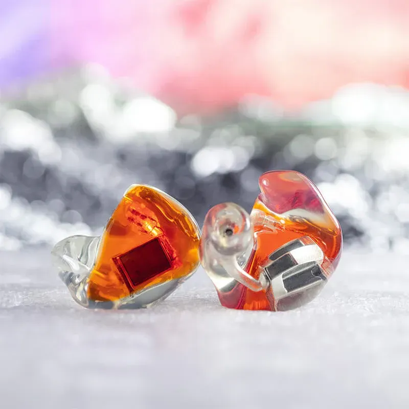 Two custom-molded, orange, semi-transparent in-ear monitor earpieces sitting side-by-side on a glittery surface.