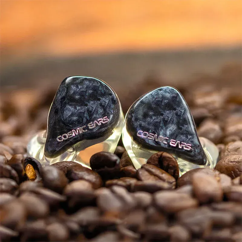 Two dark, marbled Cosmic Ears in-ear monitors resting on a bed of roasted coffee beans.