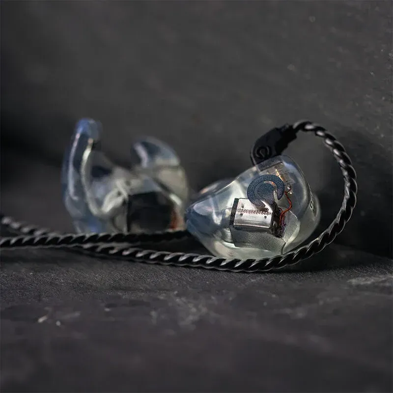 A pair of clear, custom-molded in-ear monitors with visible internal drivers, connected by a braided black cable on slate.