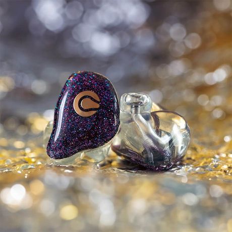 Two glittery purple wireless earbuds on a sparkling gold surface, one showing a gold “G” logo.