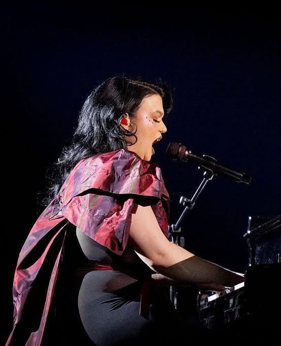 A singer called Lola Young. She has dark hair in a burgundy and black dress plays the piano while singing into a microphone