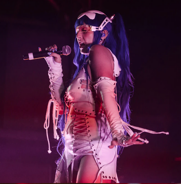 A performer with long blue hair and a futuristic white lace-up outfit sings into a microphone on a dark stage.