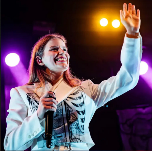 A performer in a white skeleton-print blazer smiles and waves onstage under purple and yellow stage lights.