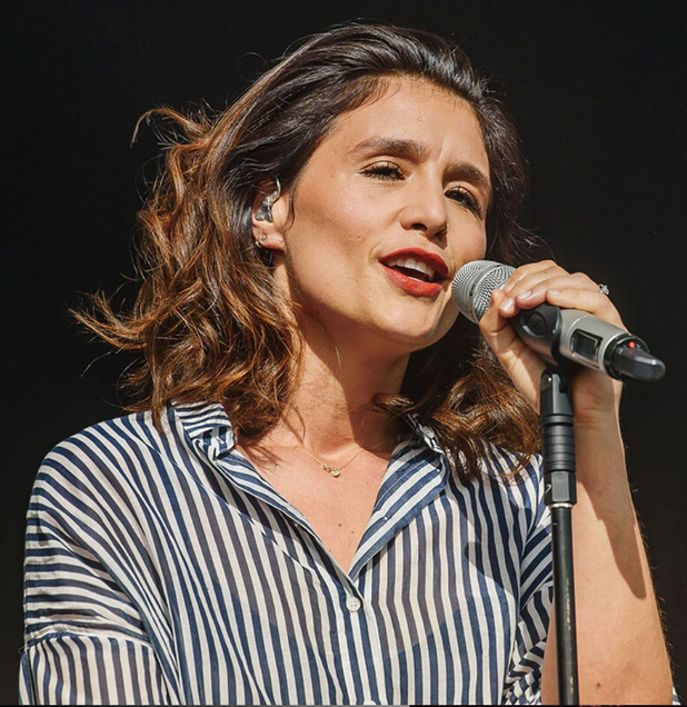 A person with shoulder-length wavy hair singing into a microphone, wearing a blue and white striped shirt.