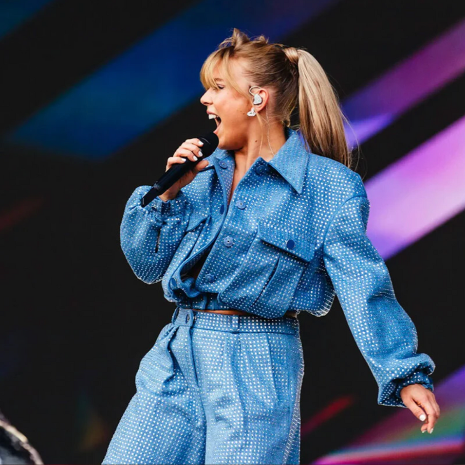 A performer singing into a microphone on stage, wearing a blue, glittery denim-style set with a high ponytail.