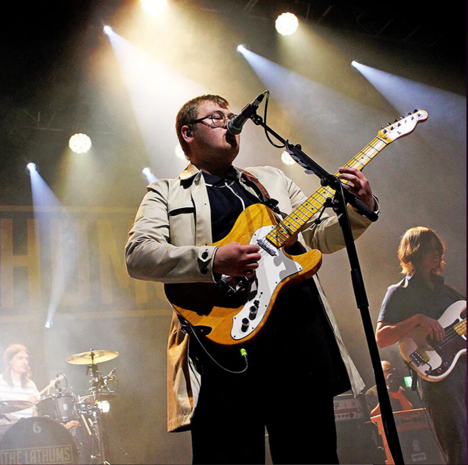 A musician singing into a microphone while playing an amber electric guitar on a brightly lit concert stage.